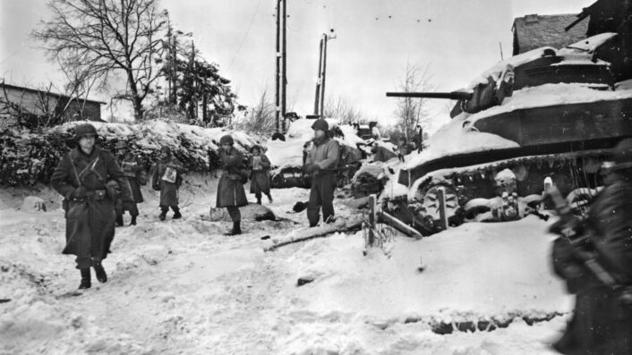 U.S. infantry pass a destroyed Stuart tank during the advance towards St. Vith, January 1945. U.S. Army photograph; public domain via Wikimedia Commons.